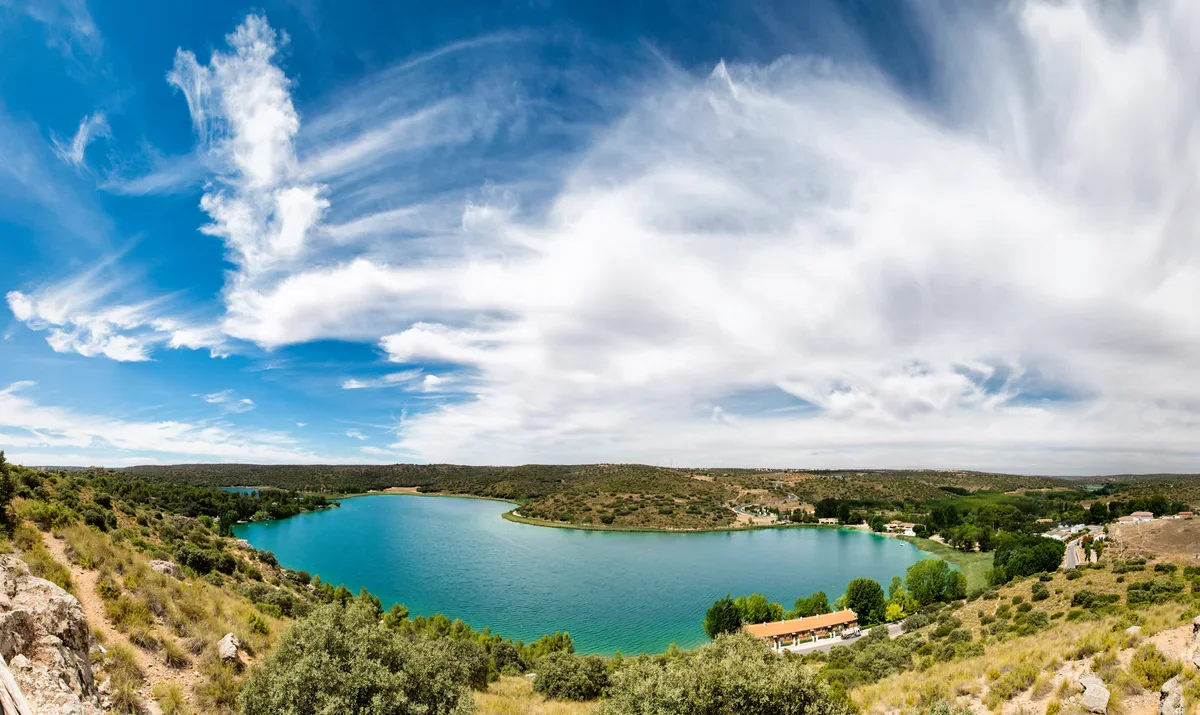 An elevated panoramic view of Laguna del Rey, surrounded by low scrub-covered hills and lakeside buildings under sweeping clouds.