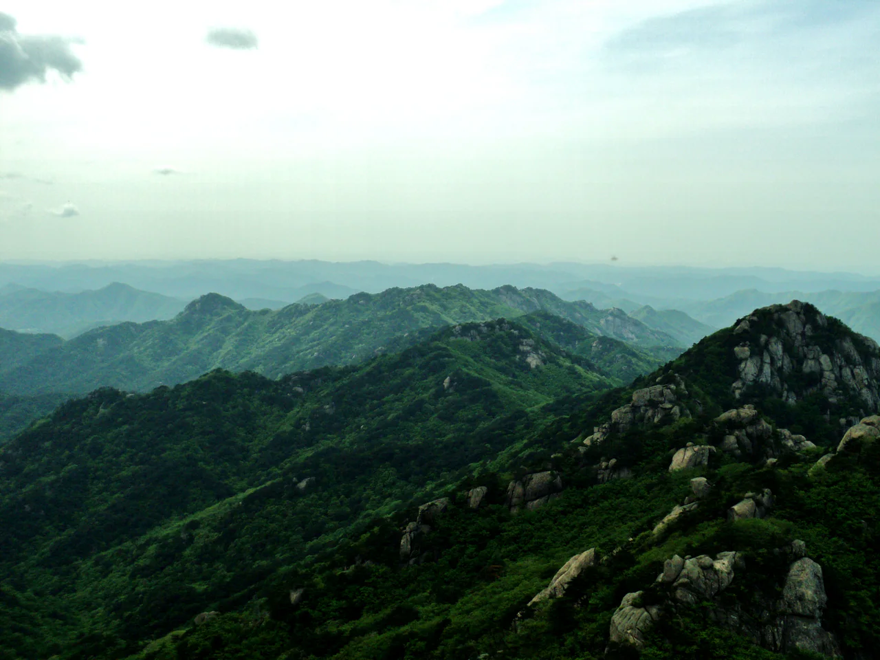Green mountain ridges and exposed rock formations recede into a hazy distance beneath a pale sky.