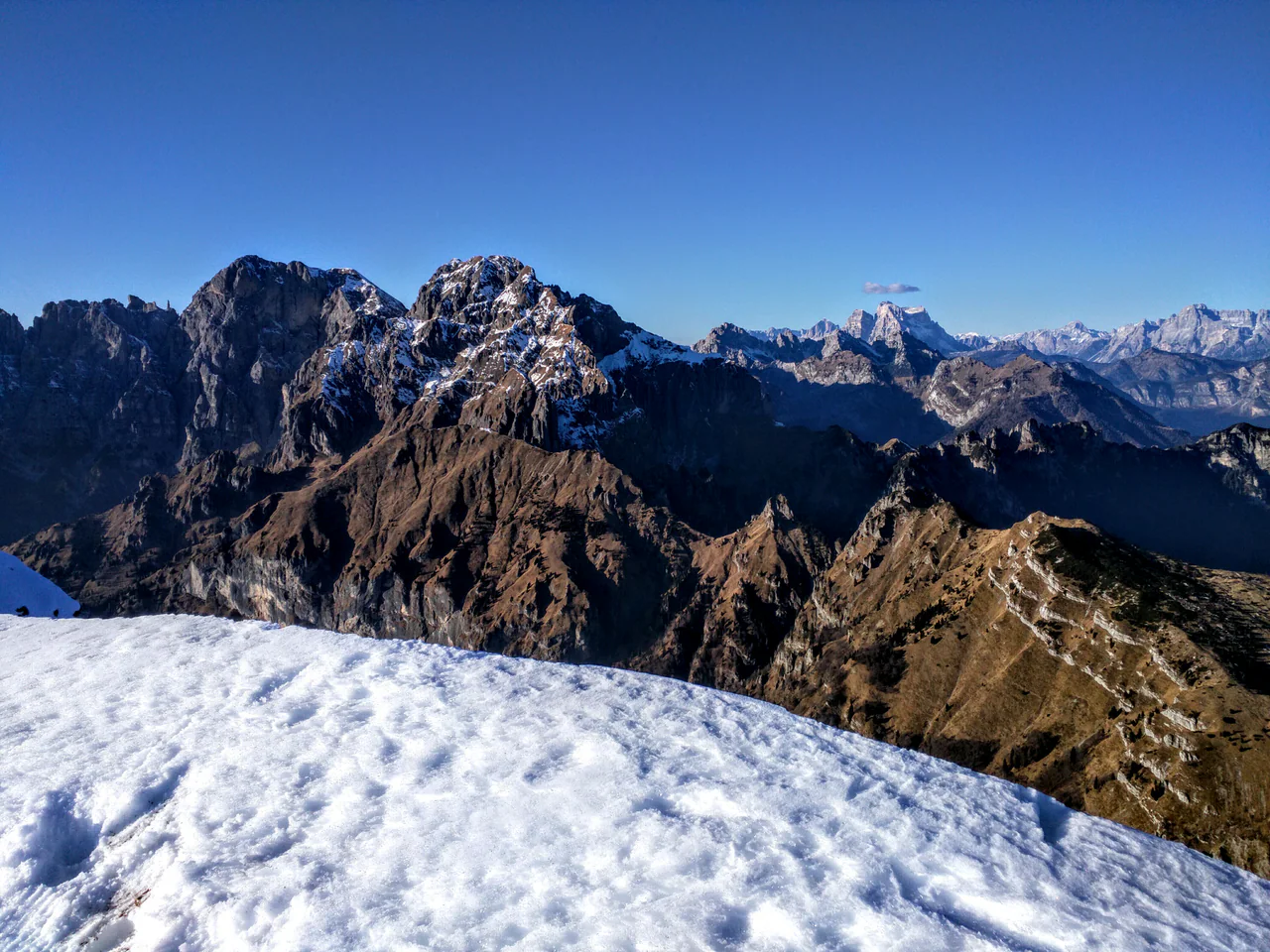 Snow covers the foreground slope, with layered grey rocky ridges and brown slopes beneath a clear blue sky.