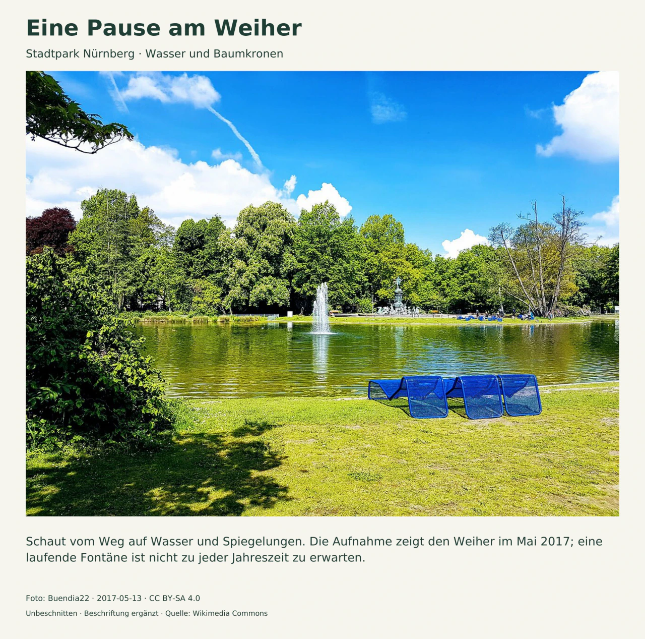 Ein Weiher mit laufender Fontäne vor dicht belaubten Bäumen; blaue Liegestühle stehen auf der Wiese im Vordergrund.