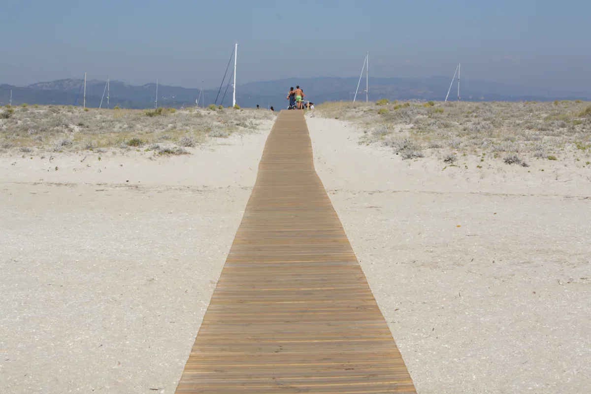 A timber boardwalk crosses pale dunes with low vegetation toward distant beachgoers and boat masts.