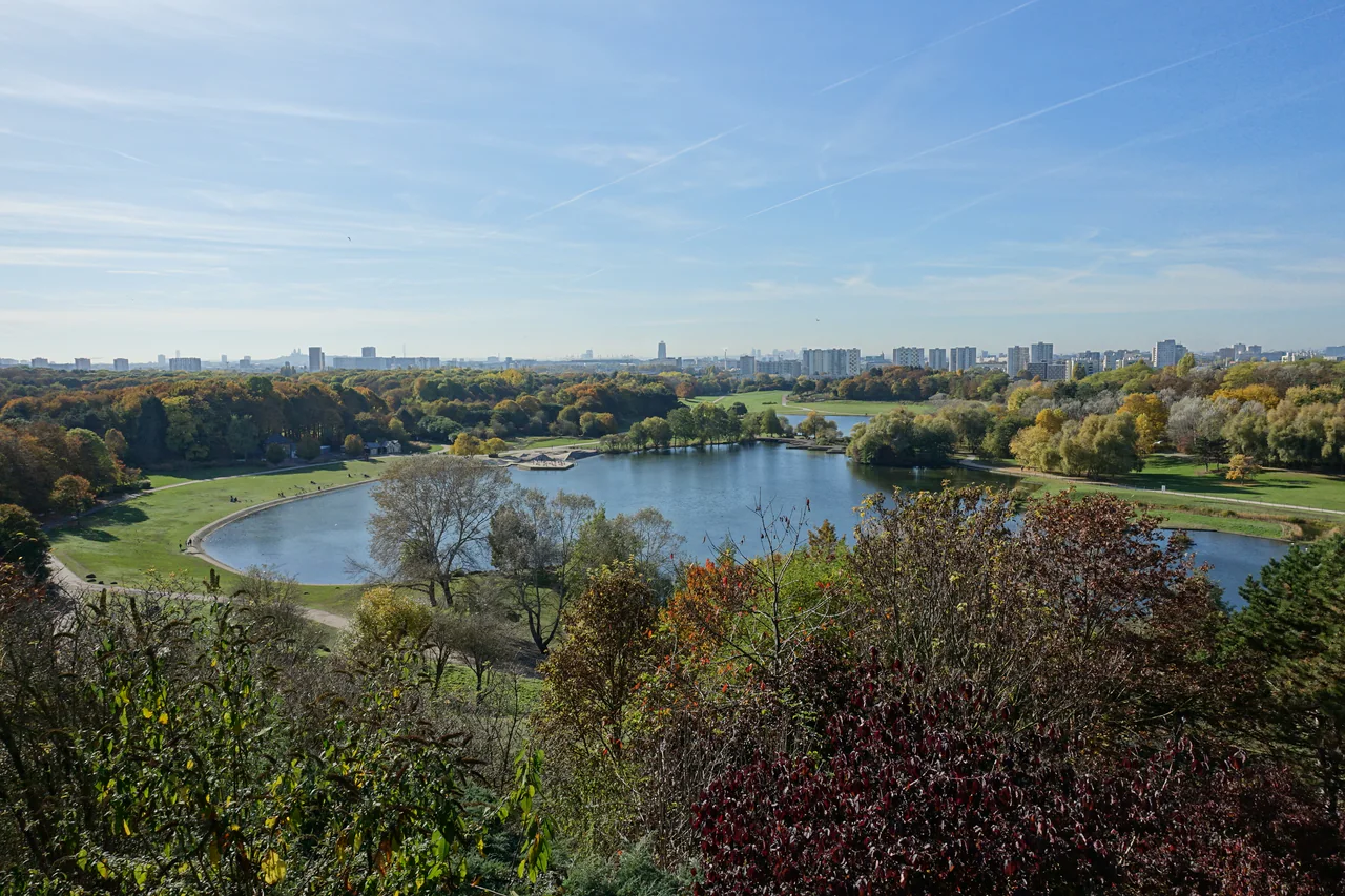 A broad lake and curving paths among autumn trees, with city buildings on the distant skyline.