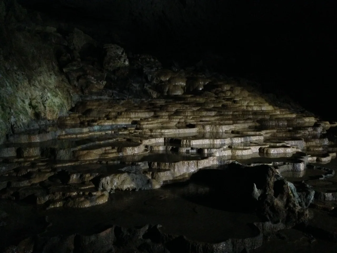 Shallow, rounded rock terraces catch the cave lighting beneath a dark cavern wall.