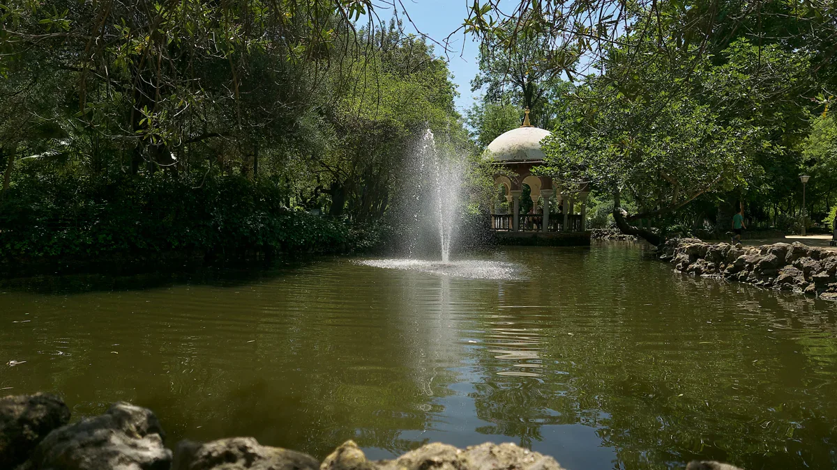 A fountain rises from a green pond in front of a small domed pavilion, framed by overhanging trees and a rocky bank.