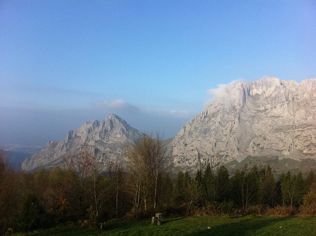 Pale limestone peaks rise beyond bare branches and evergreen woodland under a blue sky, seen from Urkiola’s Three Crosses viewpoint.