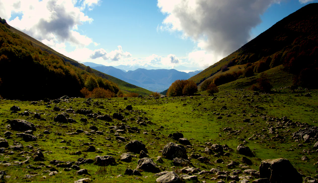 A grassy, rock-strewn valley between autumn-coloured wooded slopes, with distant mountain ridges beneath clouds and blue sky.
