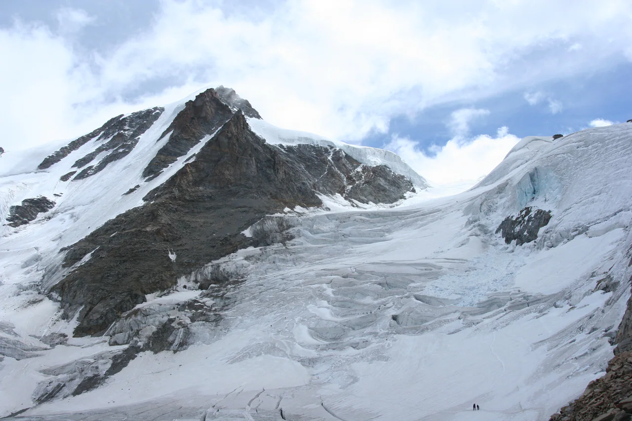 Una vetta rocciosa si alza sopra ghiaccio fratturato e neve, con minuscole figure di alpinisti vicino al bordo inferiore, in una fotografia del 2006.