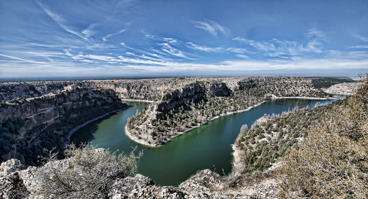 Green water curves between pale limestone cliffs and scrub-covered slopes under a wide blue sky streaked with white clouds.