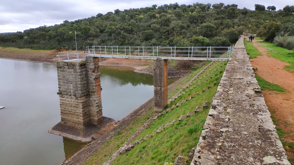 A stone water-intake tower linked by a metal walkway to the dam beside Cornalvo reservoir, with wooded slopes behind.