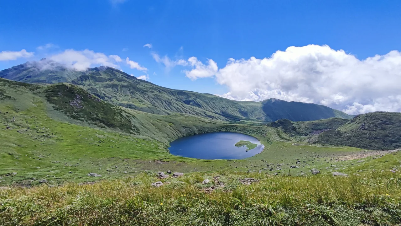 A deep blue lake sits in a hollow among green mountain slopes, with a rocky ridge, white clouds and blue sky beyond.