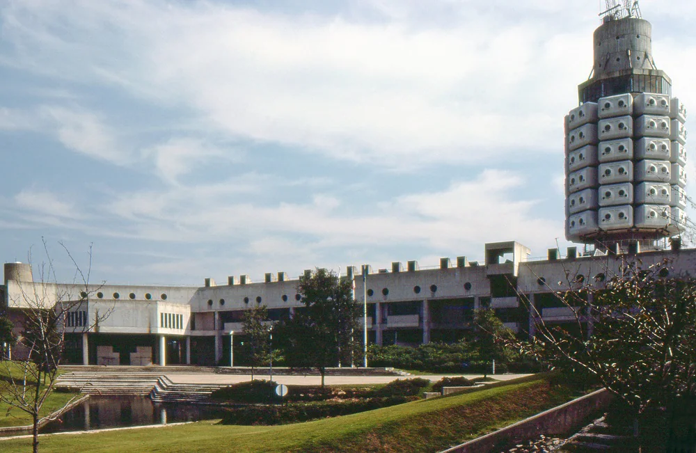 A low concrete building stands behind a shallow water feature and lawn, with a tall pod-like tower at the right.