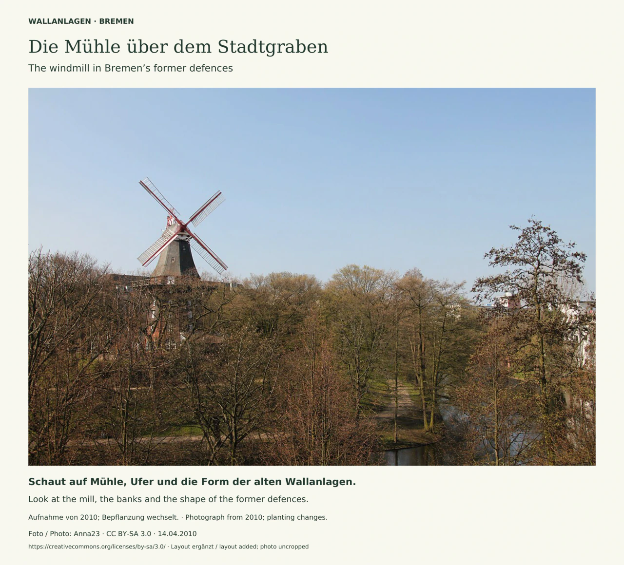 Eine Windmühle mit vier weiß-roten Flügeln ragt links über noch weitgehend kahle Bäume. Rechts unten windet sich der Stadtgraben zwischen baumbestandenen Ufern; der Himmel ist klarblau. Zweisprachiger Titel, Bildhinweis und Fotocredit rahmen das Foto.