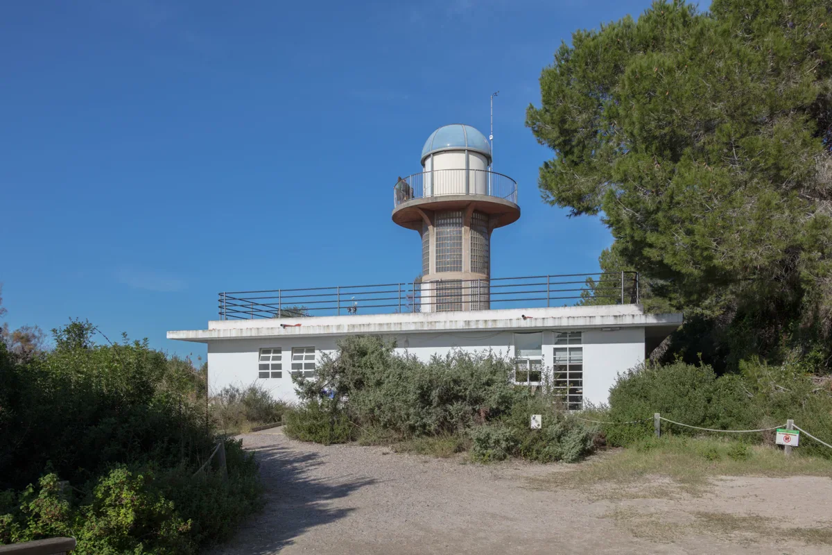 A white observation building with a raised round tower stands beside shrubs and tall trees, with an earth path in front.