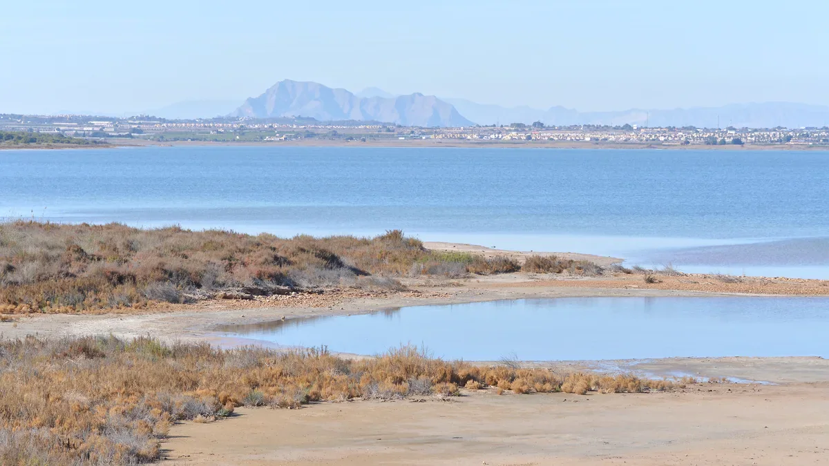 Blue lagoon water surrounds a low spit edged with scrub, with buildings and pale mountain ridges across the water.