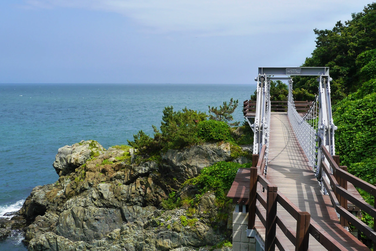 An empty suspension footbridge and wooden approach stand beside a rocky, green coastal slope and open blue sea at Igidae.