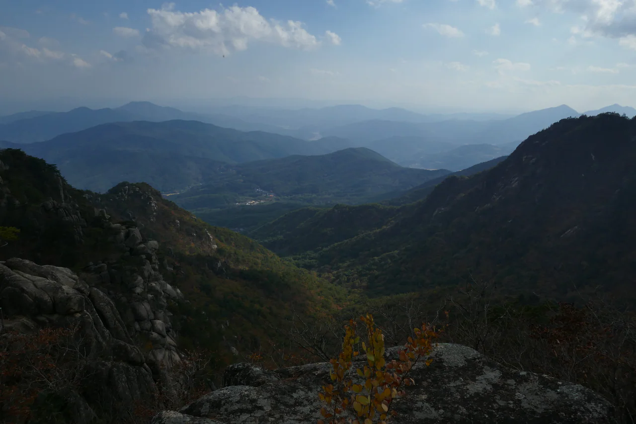 Granite rocks and autumn-tinted forest in the foreground, with layered blue mountain ridges beneath a cloudy sky.
