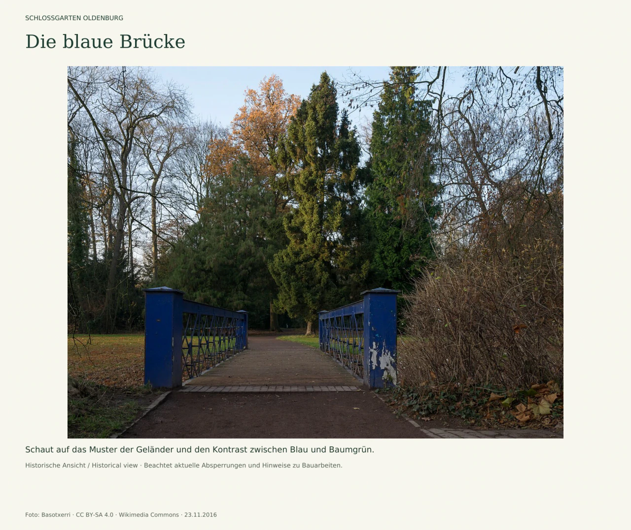 Eine kleine Brücke mit blauen Pfosten und rautenförmigen Eisengeländern führt geradewegs auf einen Parkweg. Dahinter stehen hohe immergrüne und teils kahle Bäume; Laub liegt am Wegrand.