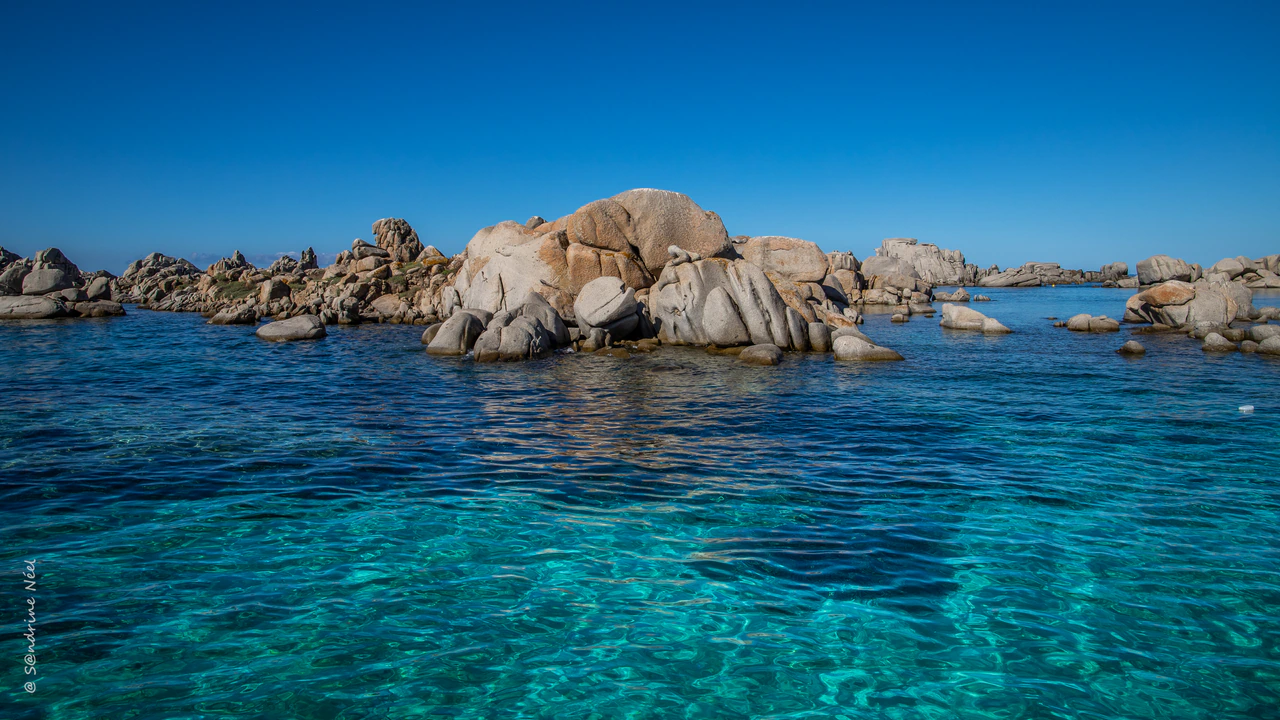 Rounded pale granite islets rise above blue and turquoise sea under a clear sky; a vertical photographer’s signature remains at lower left and a small white object floats at right.
