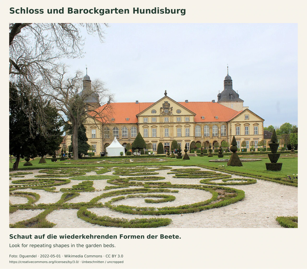 Geschwungene, niedrige Beetumrandungen auf hellem Kies vor der gelben Schlossfassade mit rotem Dach und zwei Türmen. Geschnittene Gehölzformen stehen auf dem Rasen; links ragen noch kahle Äste ins Bild.
