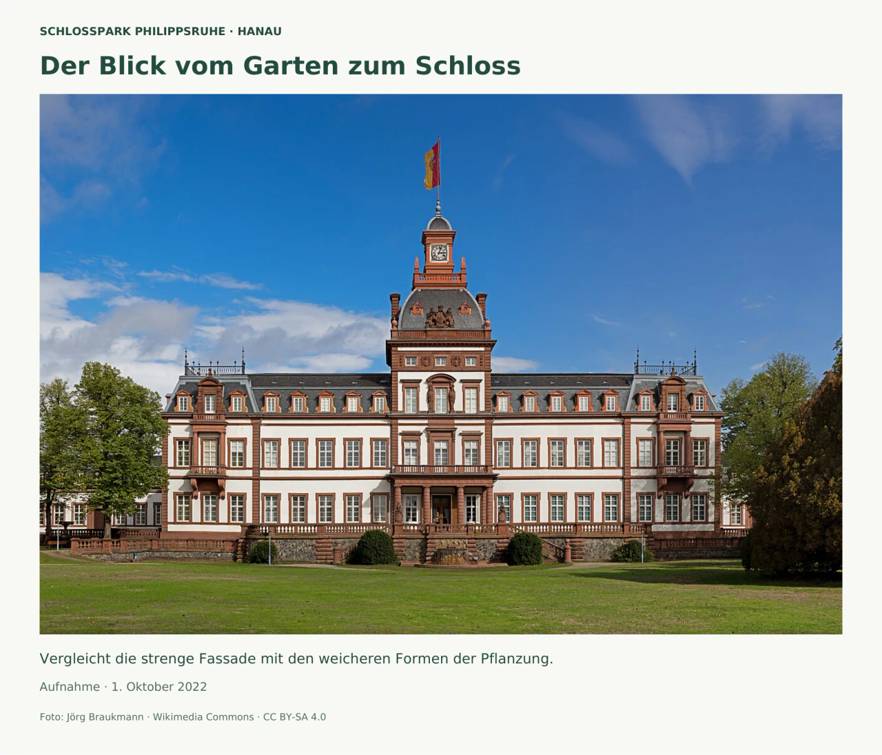 Frontaler Blick über eine Rasenfläche auf die weiße, rot gegliederte Schlossfassade mit symmetrischen Fensterreihen, einem mittigen Uhrturm und Fahne. Bäume rahmen die Seiten unter blauem Himmel.