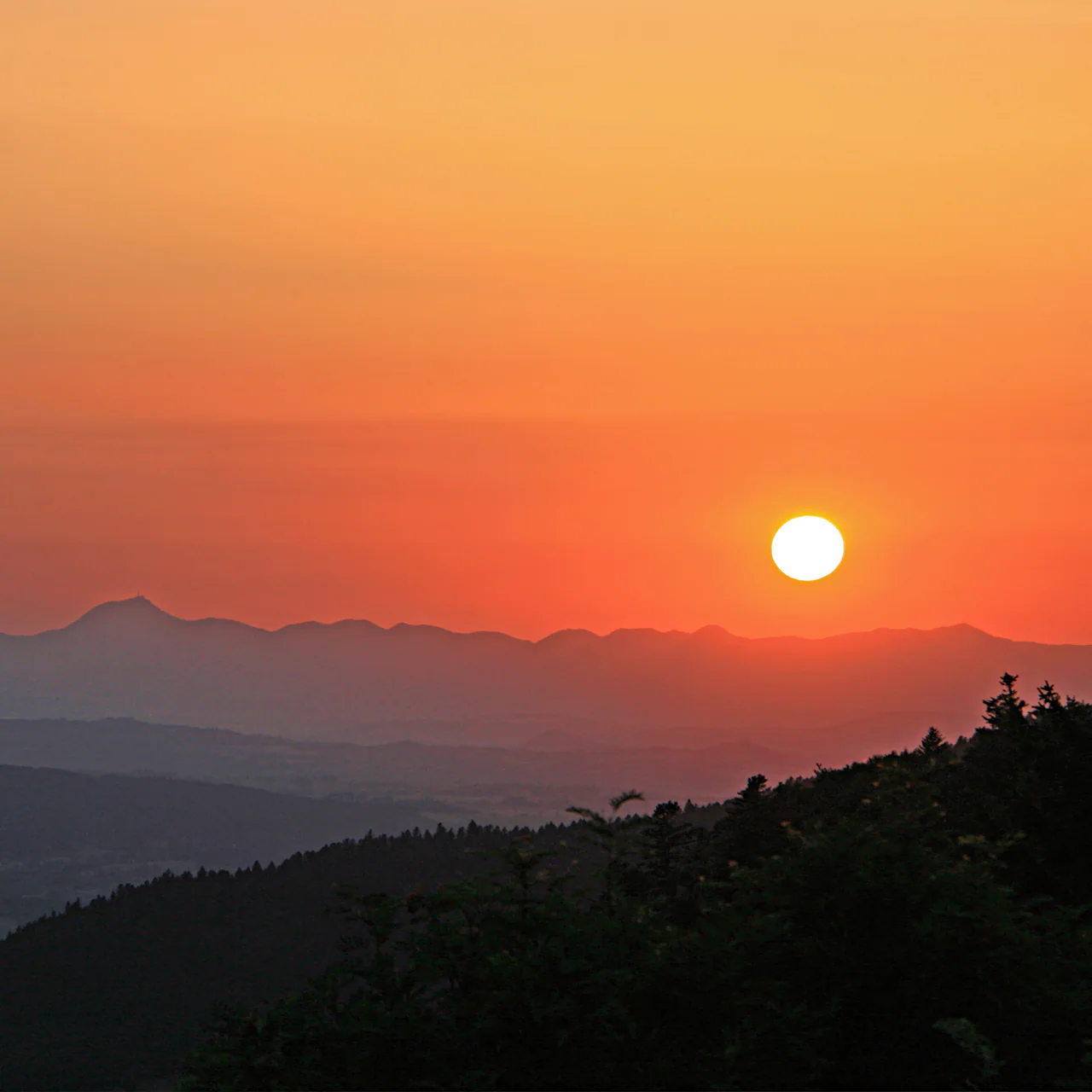 Bright evening sun above layered dark mountain ridges under an orange sky, with wooded slopes in the foreground.