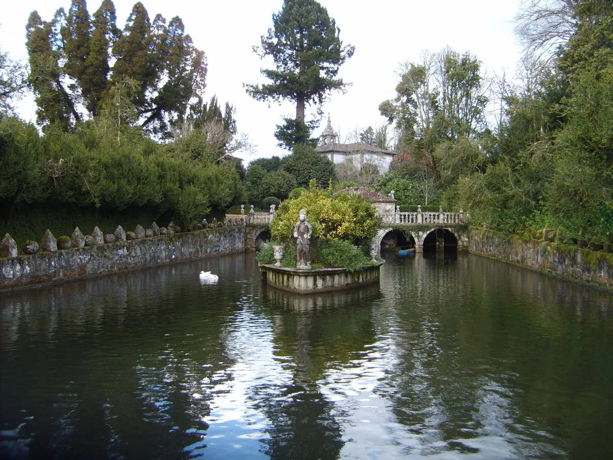 A stone-edged pond surrounds a planted ornamental island with a statue; an arched stone bridge and tall trees stand behind it.
