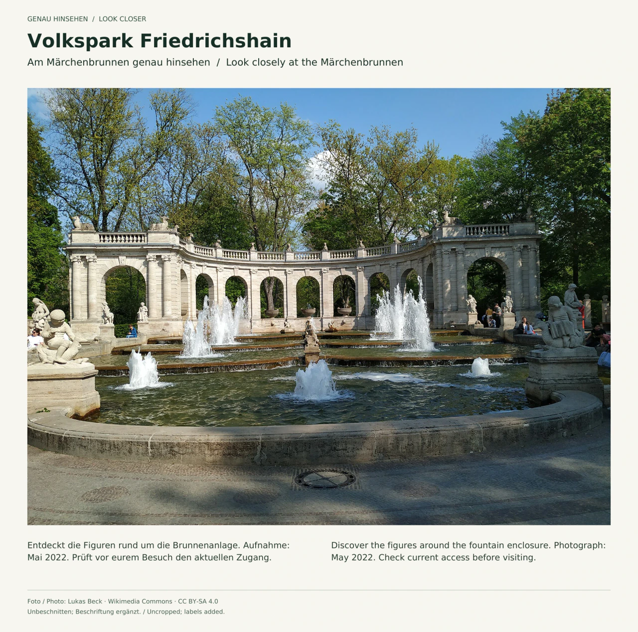 Der Märchenbrunnen im Volkspark Friedrichshain im Mai 2022: mehrere Wasserfontänen in gestuften Becken, steinerne Märchenfiguren am Rand und eine gebogene Arkadenwand vor Bäumen; einige Besucher sitzen am Rand.