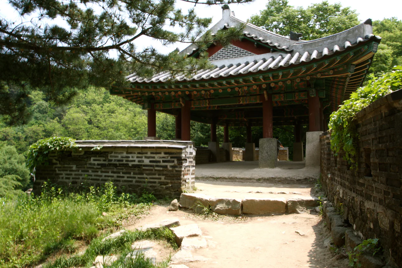 A tiled roof with painted wooden eaves stands above stone walls and an uneven foreground, framed by green trees.