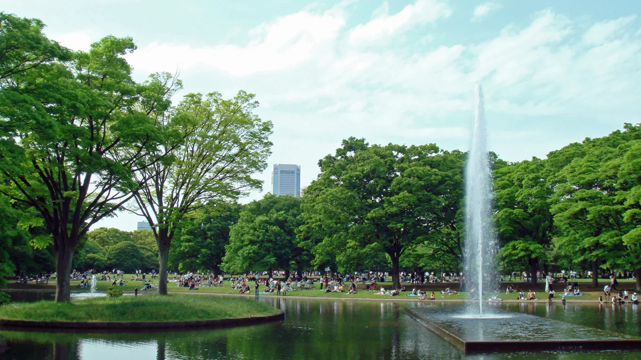 A tall fountain rises from a pond beside leafy trees and lawns where visitors are relaxing.