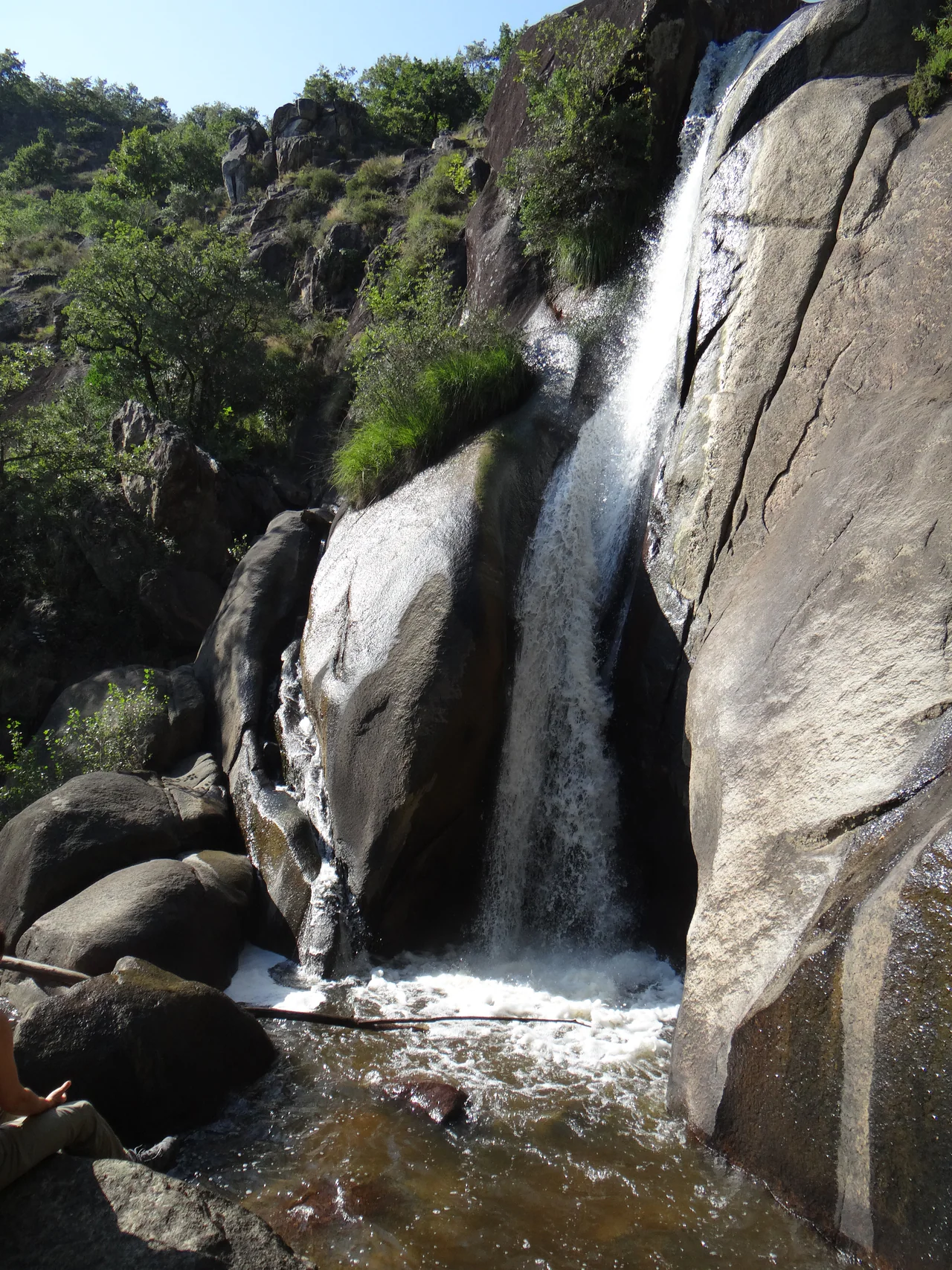 A waterfall runs down tall rounded grey rock into a brown pool, with vegetation above and part of a seated person visible at the lower left.