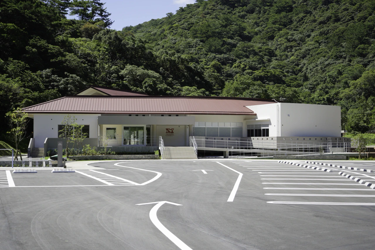 A white visitor-centre building with a red roof and entrance ramp stands in front of a densely wooded hillside, beyond an empty parking area.