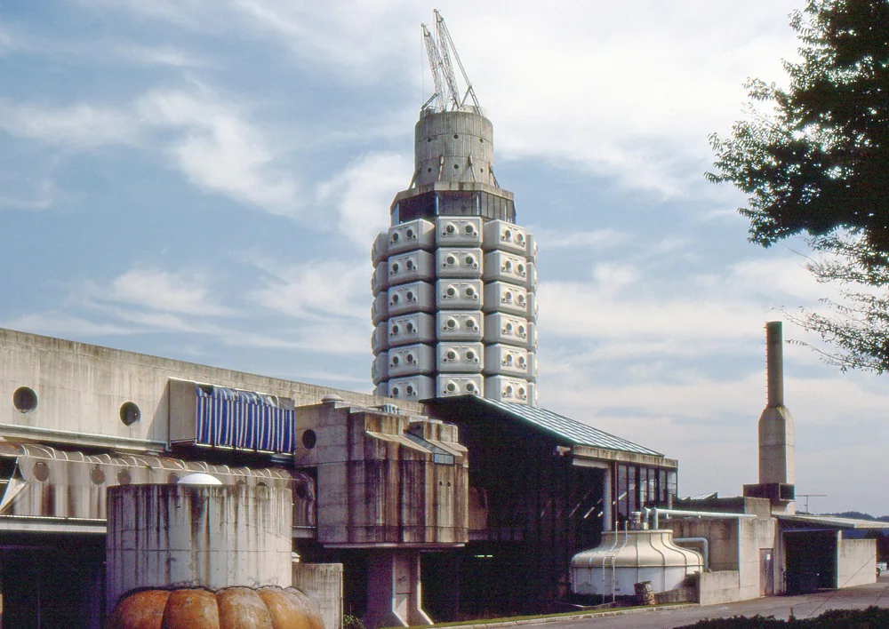 A tower of stacked rounded modules rises above angular concrete buildings and a sloping roof, with a tree at the right edge.