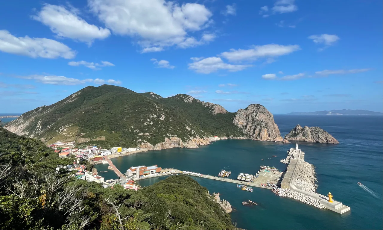 Blue water surrounds a Hongdo harbour with breakwaters, village buildings and a wooded rocky ridge behind it.
