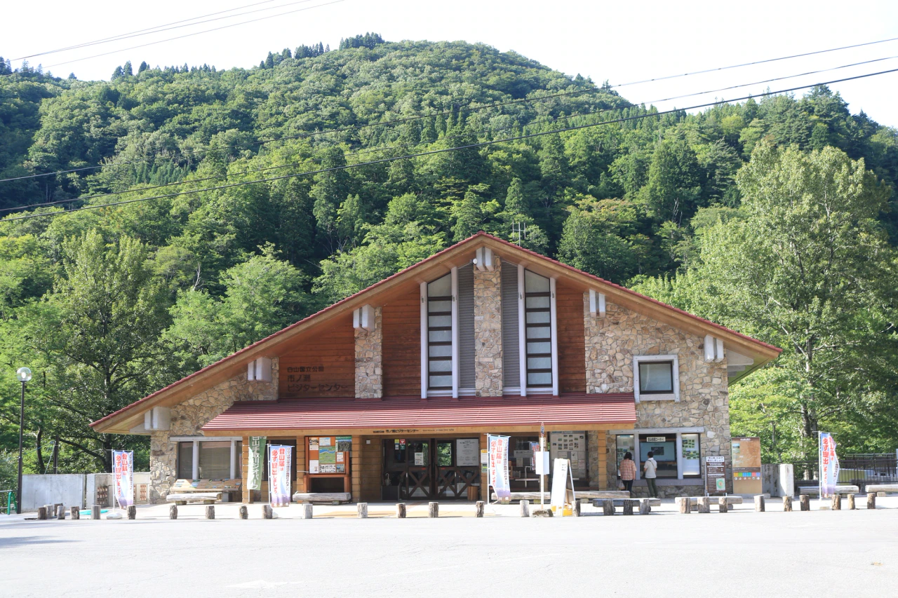 A broad timber-and-stone visitor-center building with a red pitched roof stands below a green wooded hillside, with notices around its entrance.