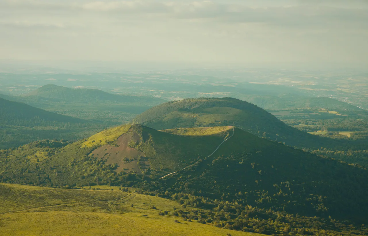 A green volcanic cone with a circular crater rises among wooded hills under a hazy sky.