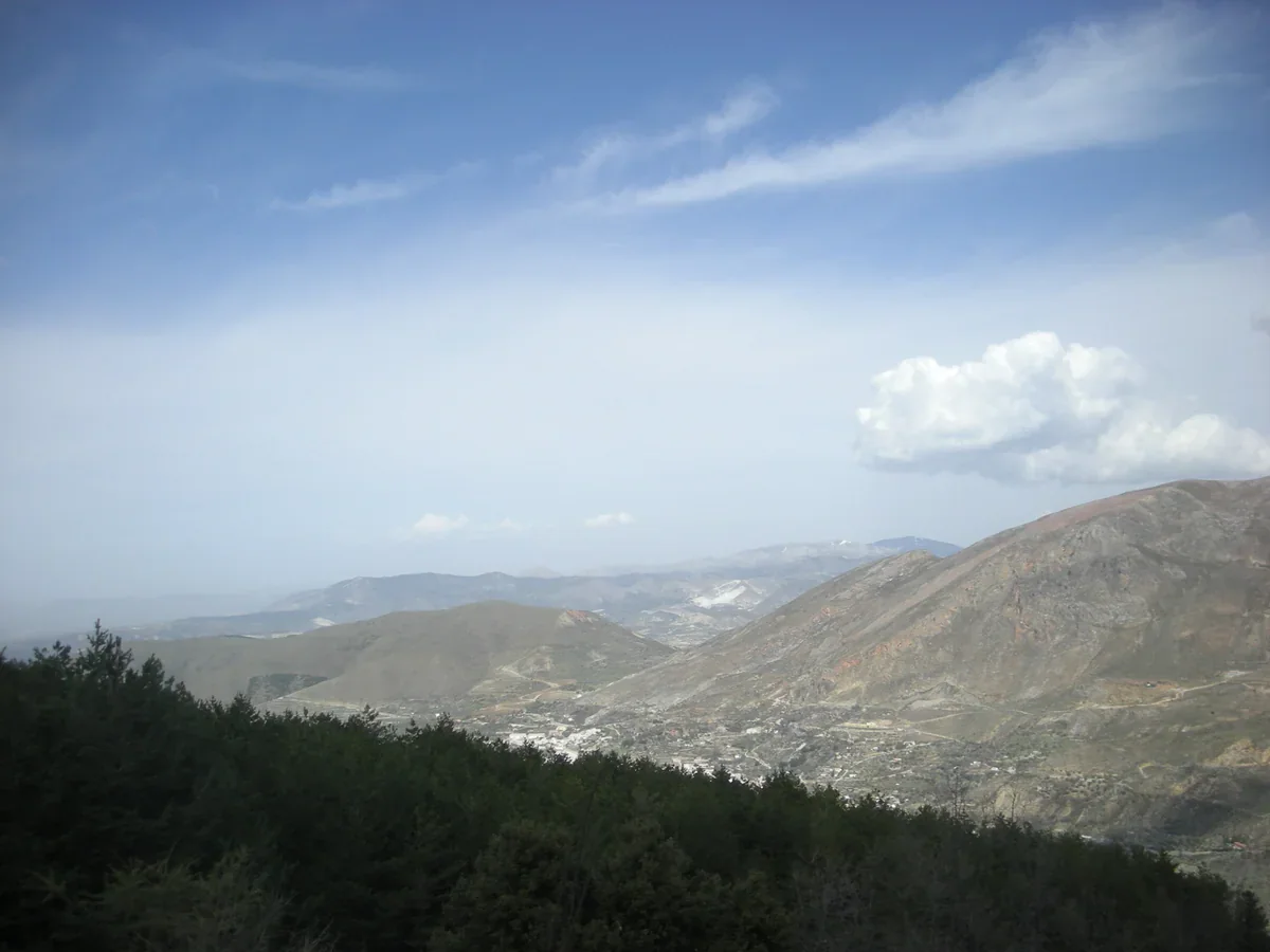 Pine-covered foreground slopes overlook a hazy mountain valley with scattered buildings under streaked clouds.